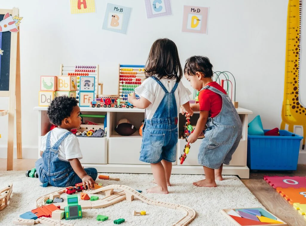 Two toddlers in denim overalls playing with toys and a wooden train