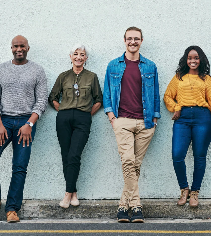 Four friends of different ages smiling while leaning against a pale wall
